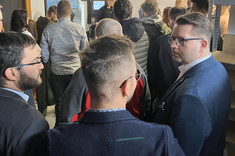 Grupa uczestników w hali produkcyjnej, niektórzy wymieniają uwagi, inni obserwują prezentację na monitorze 